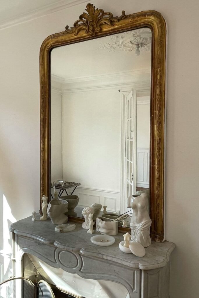a styled vignette with a large gold-framed mirror above a white vintage dresser, with candles or perfume bottles. styled as Parisian bedroom ideas
