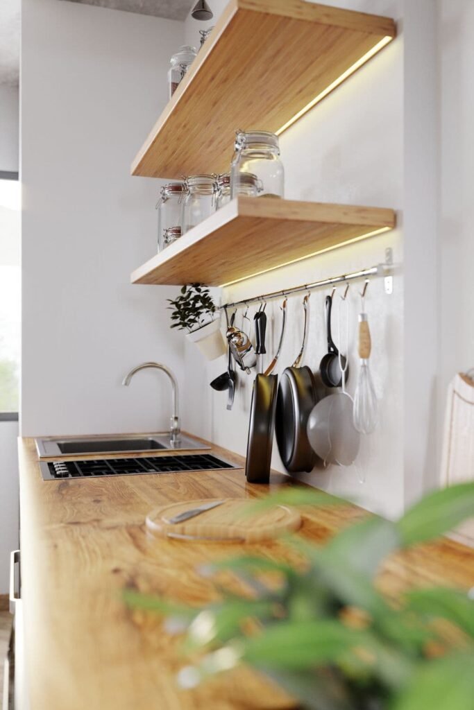 A small apartment kitchen with pots and pans hanging from ceiling racks above a kitchen island, surrounded by cabinets, shelves, and a window letting in natural light.