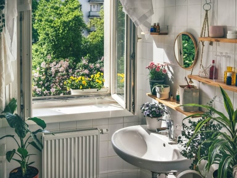Small apartment bathroom with a cozy, plant-filled decor setup. Features include a white ceramic sink, wooden floating shelves, and low-maintenance indoor plants like spider plant, carnations, and purple flowers. Natural sunlight streams through an open window, enhancing the biophilic, modern aesthetic. Perfect inspiration for cozy small apartment bathroom decor ideas, modern small bathrooms with plants, and space-saving styling for renters.
