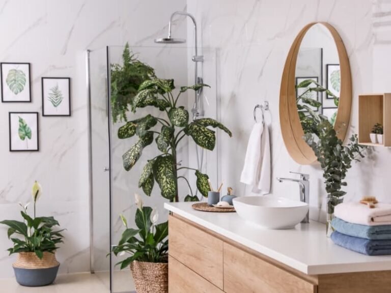 Small apartment bathroom with a cozy, plant-filled decor setup. Features include a white ceramic sink, wooden floating shelves, and low-maintenance indoor plants like spider plant, carnations, and purple flowers. Natural sunlight streams through an open window, enhancing the biophilic, modern aesthetic. Perfect inspiration for cozy small apartment bathroom decor ideas, modern small bathrooms with plants, and space-saving styling for renters.