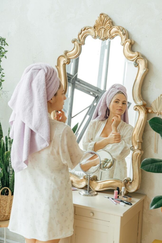 A young woman stands in front of an ornate gold-framed mirror, wearing a light floral robe and a lavender towel wrapped around her hair. She gently touches her face while looking at her reflection. The vanity table in front of her holds makeup items including brushes, lipstick, and nail polish. The room has a soft, natural aesthetic with green plants and soft lighting, creating a serene and cozy atmosphere.
